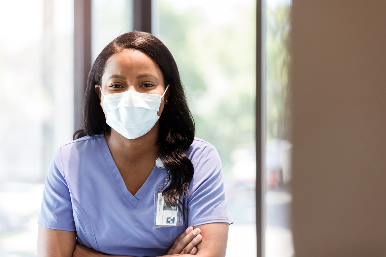 A confident mid adult female nurse wearing a protective face mask looks at the camera. She is standing with her arms crossed.
