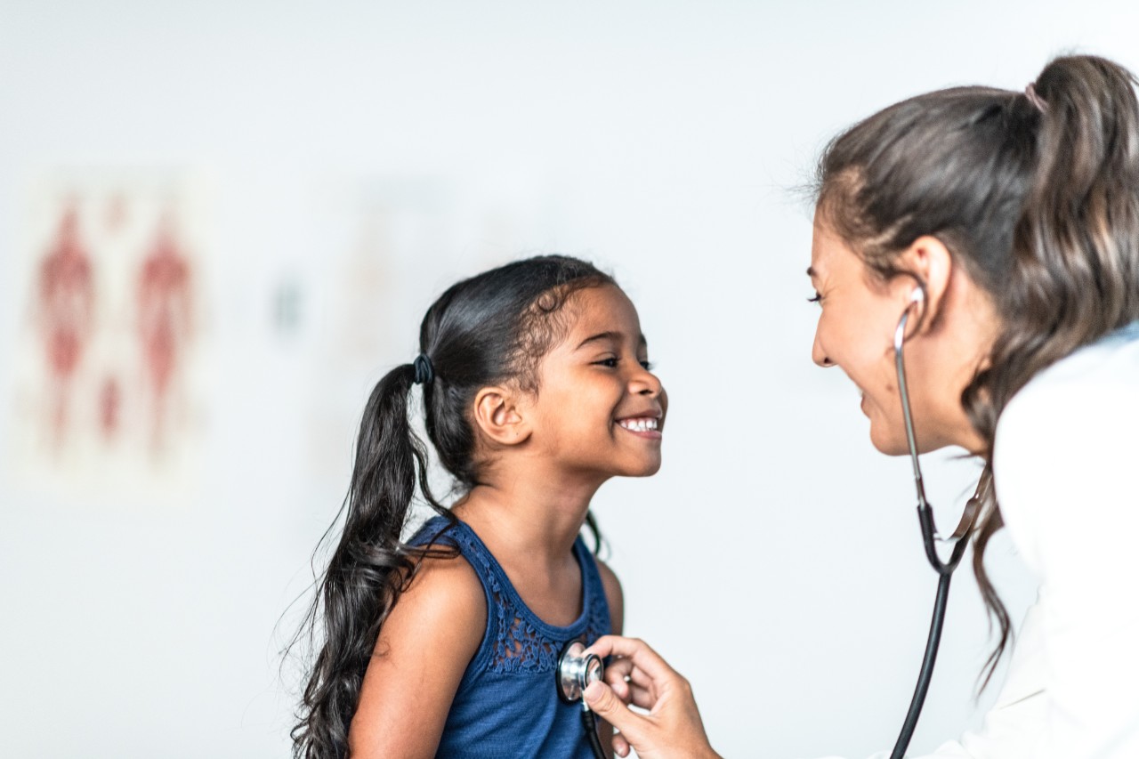 A young East Indian girl is examined by her Hispanic female doctor.  The doctor is checking her heart with the stethoscope that is around her neck.  The young girl is wearing a blue tank top while the doctor is wearing a white lab coat.