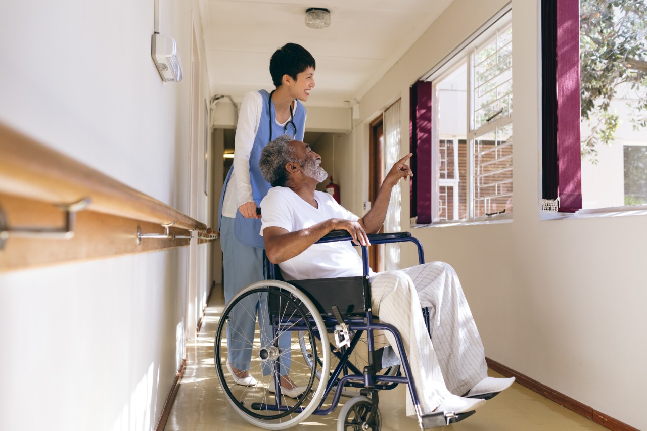 Side view of  Asian female nurse and senior African-american male patient interacting with each other while standing in corridor at retirement home